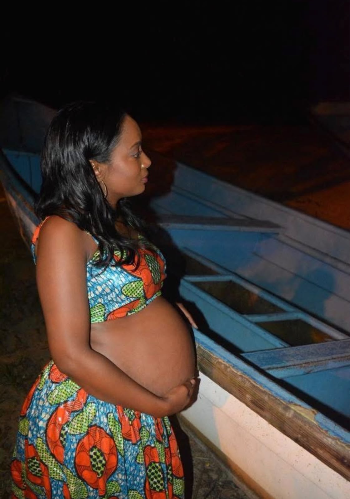 Woman in vibrant African print standing beside a fishing pirogue at night on the Moruga coast